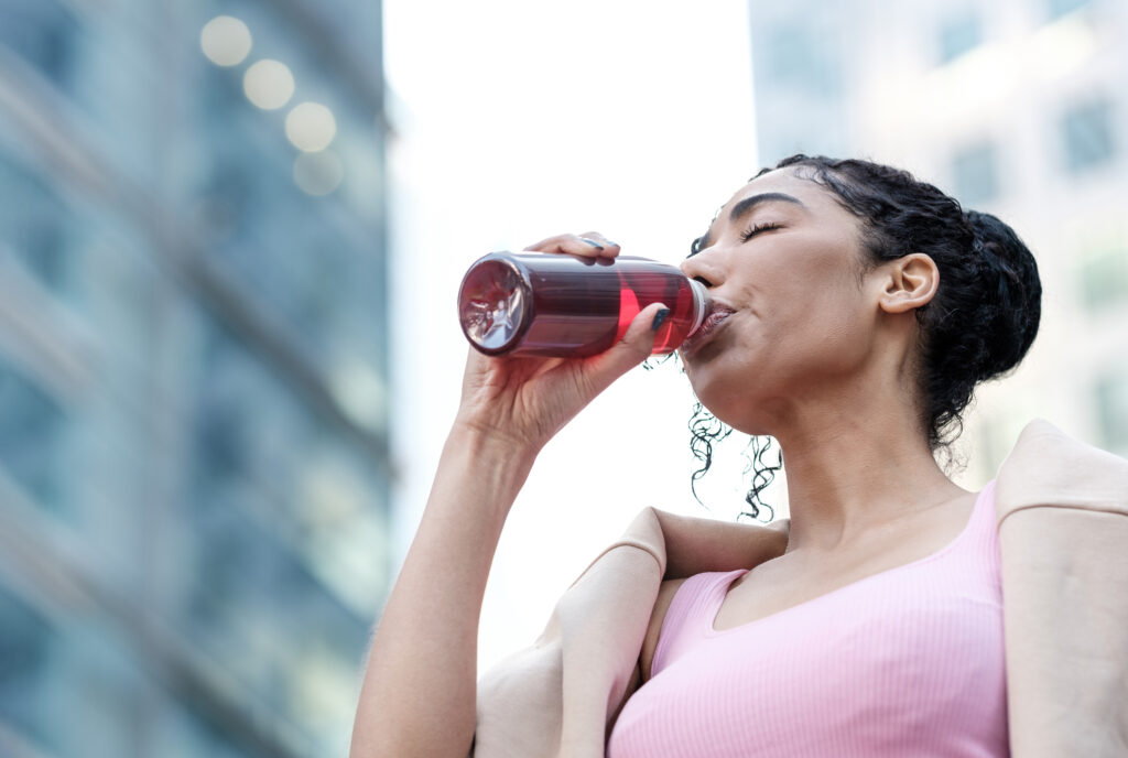 Portrait of young fitness woman drinking energy drink from a bottle. She has her eyes closed and she is wearing a pink sport bra and a sweater on her shoulders. Fitness and hydration concept. Healthy lifestyle.