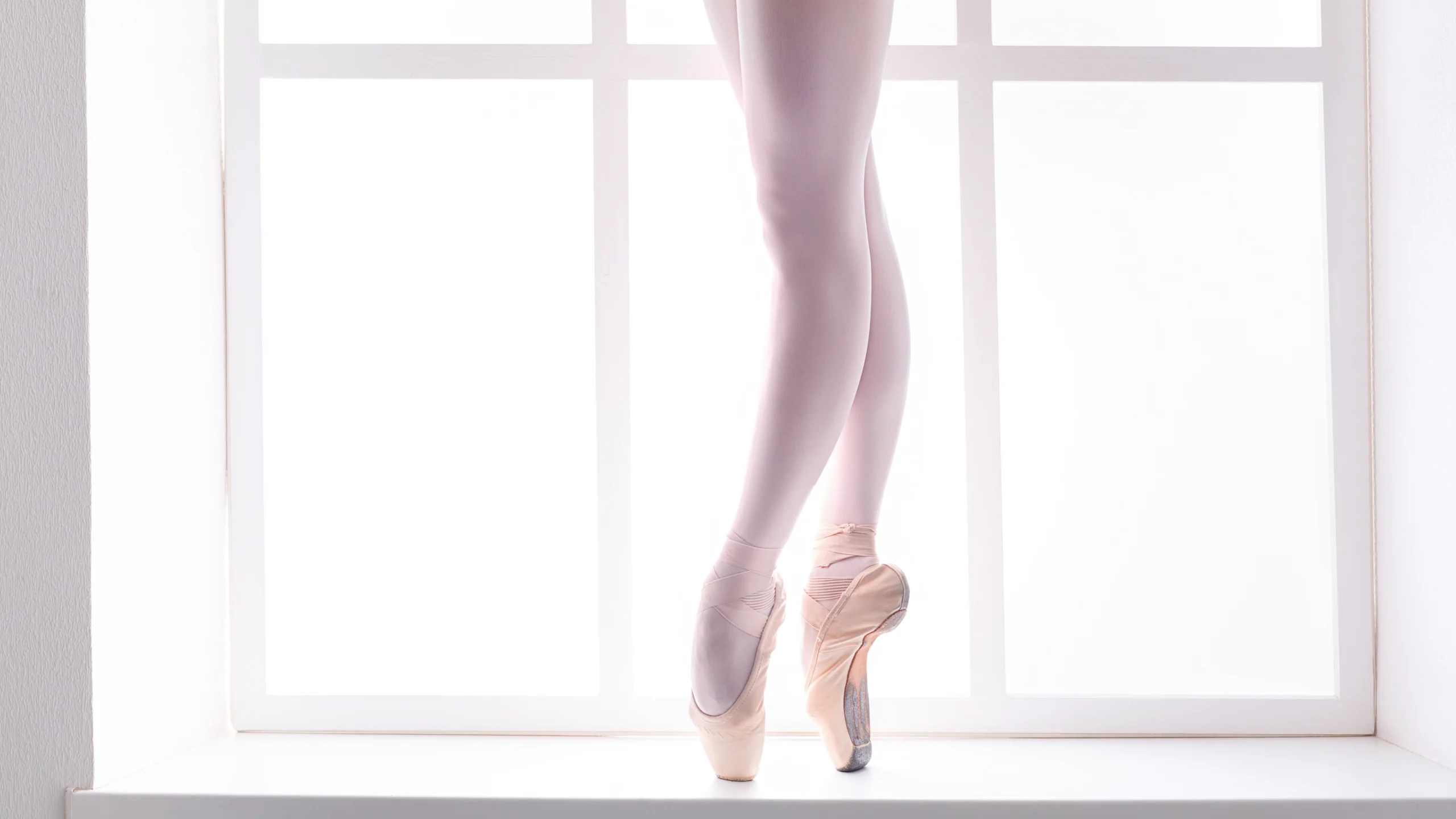 A female dancer with hyperextended knees is shown from the hips down posed in sus-sous croisé on pointe, framed by a large window. She wears a black leotard, pink tights and pink pointe shoes.
