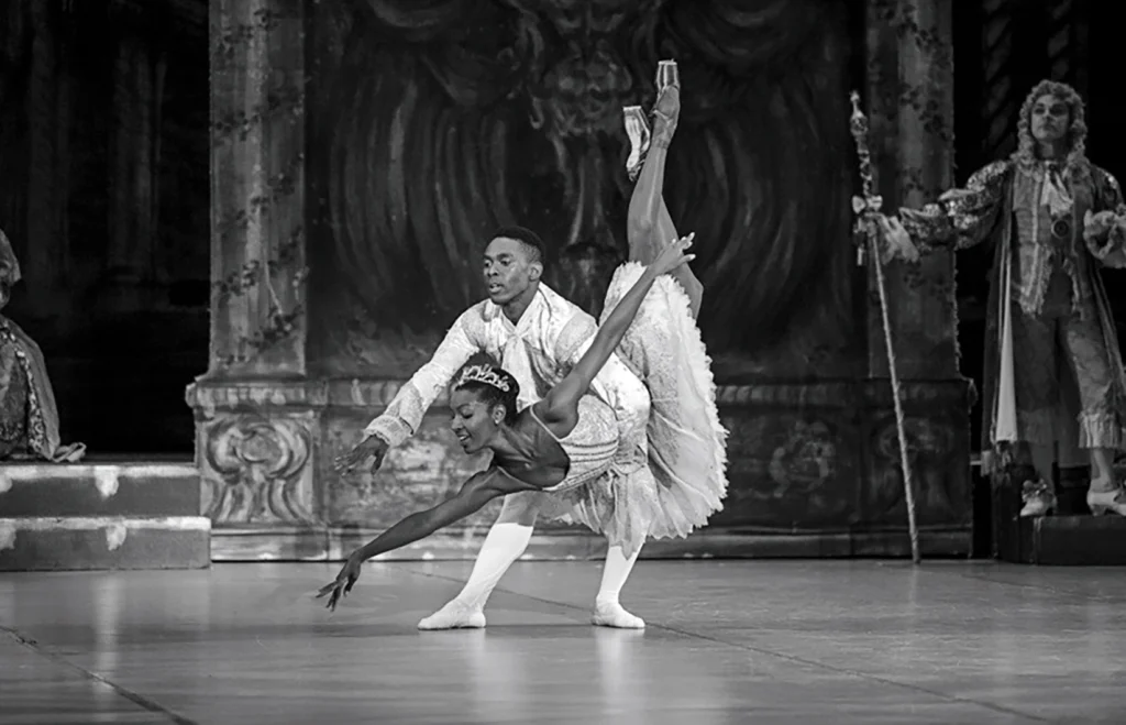 In this black and white photo, Andile Ndlovu leans forward and holds Precious Adams by the waist as she plunges into a fish dive. They are performing onstage in the ballet The Sleeping Beauty, with Adams wearing a light tutu and crown and dark tights and pointe shoes, and Ndlovu in a white tunic tights and ballet slippers.