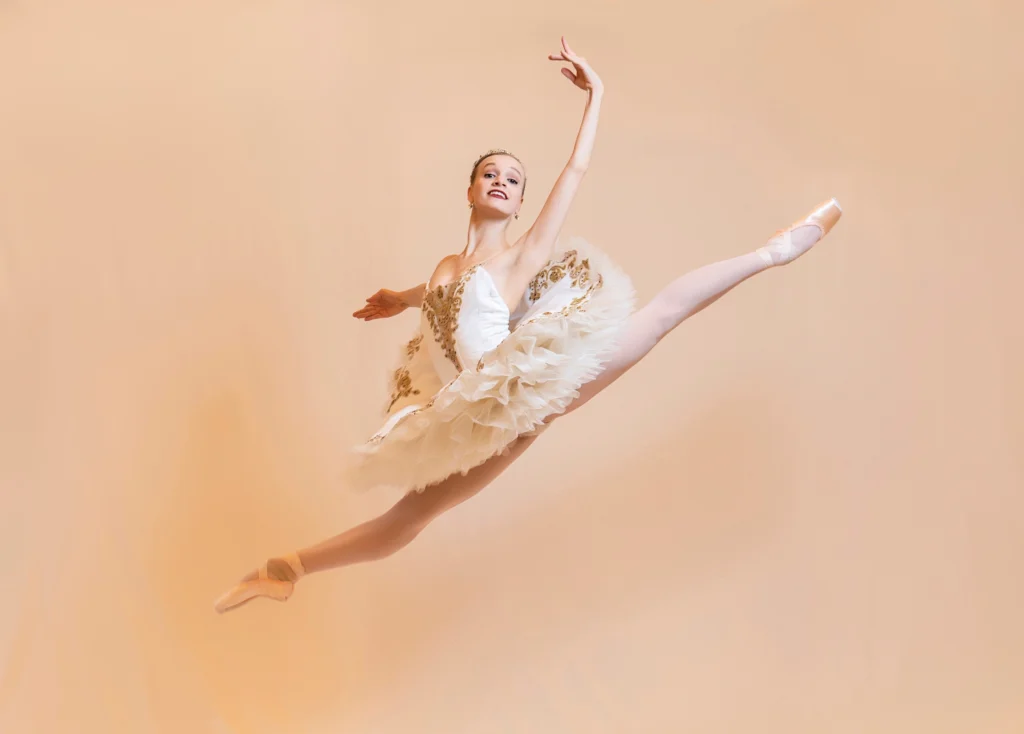 Jennifer Hackbarth poses in a soaring saute in a perfect split, arms in high third. She wears a white and gold tutu, and the background is a light peach color. 