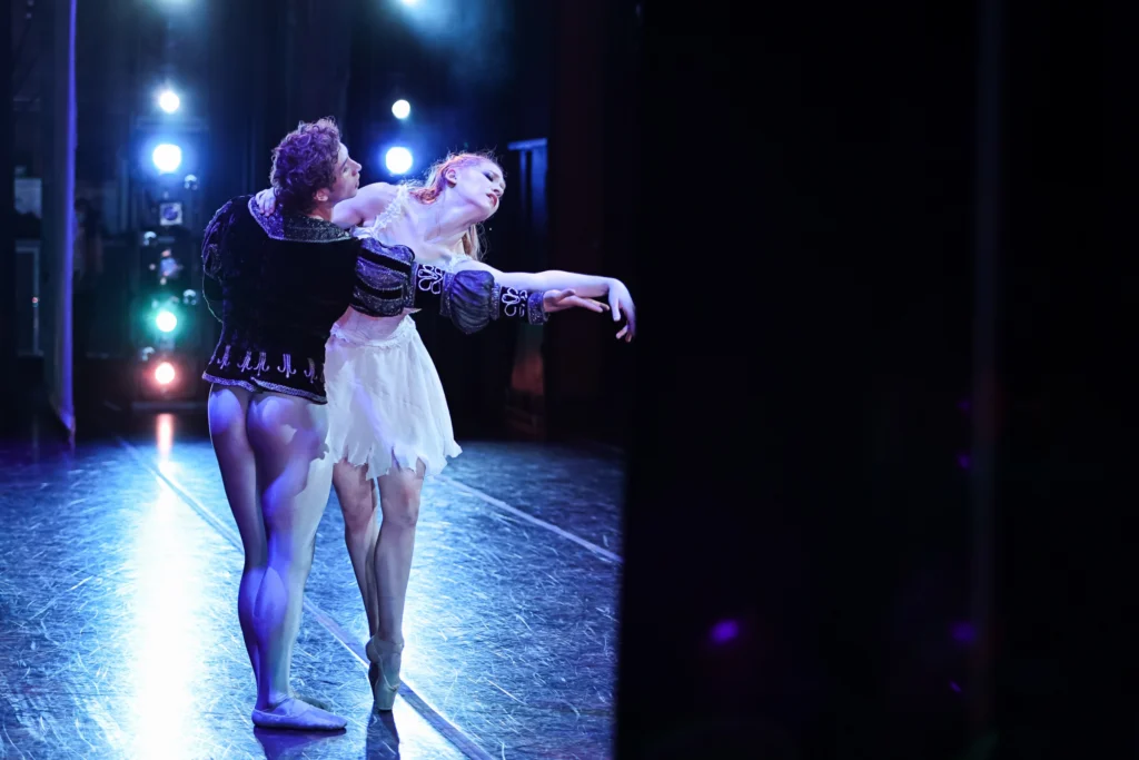 Lizzie Tripp and Parker Brasser Vos dance together onstage, the photo capturing them from the wings. Tripp reaches offstage on point, Brasser Vos reaching for her arm and pulling her toward him.