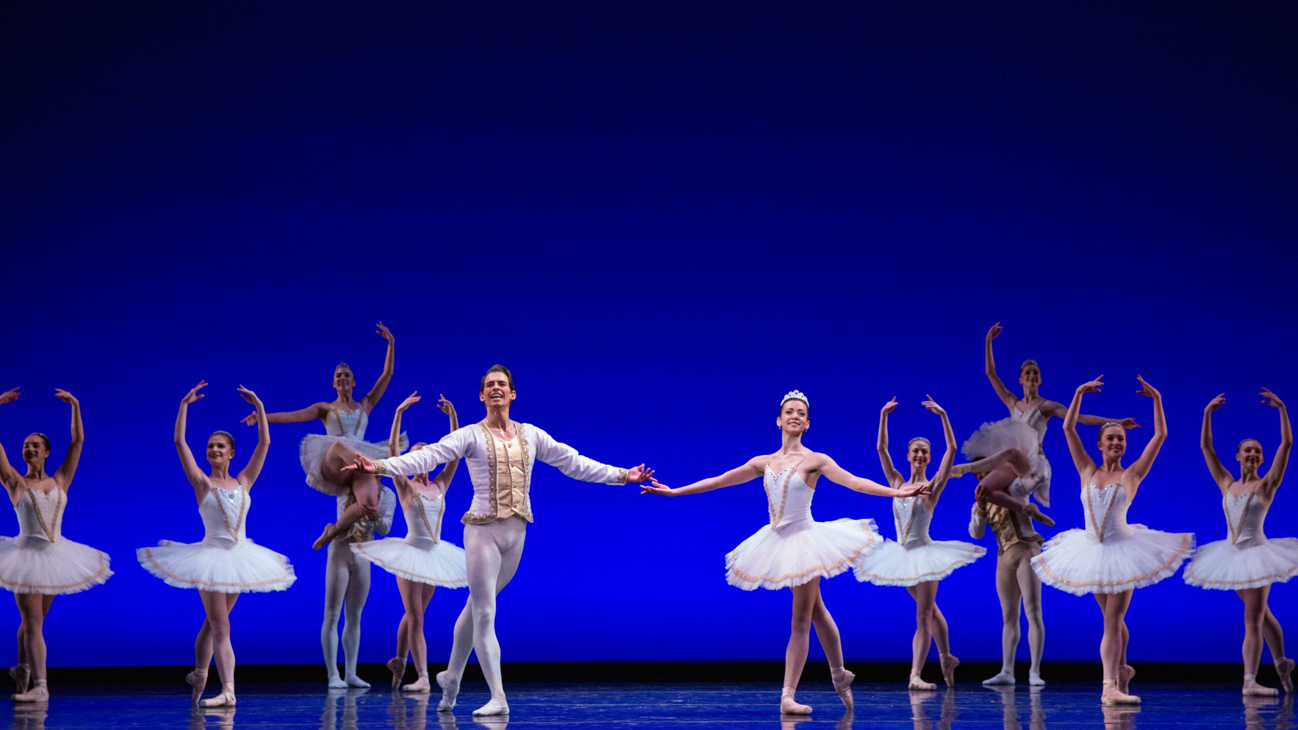 Onstage in front of a deep blue backdrop, an ensemble of dancers pose in tendu derriere en croisé, the men in white tunics and tights, and the women in white tutus.