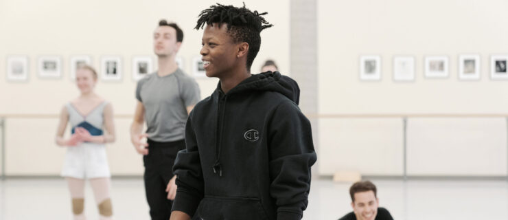 Siphesihle November is shown from the thighs up in a large dance studio, smiling at someone off camera to his right. He wears a black hooded sweatshirt and black sweatpants. Four other dancers, two women and two men, stand or sit behind in, smiling and laughing.