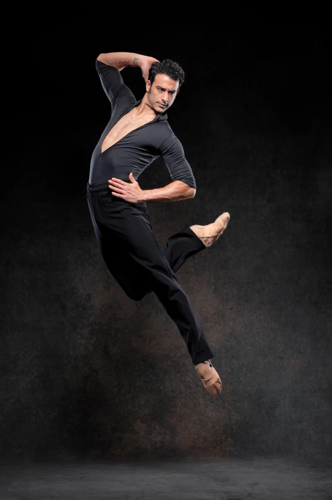Arian Molina Soca jumps high, tucking his right leg back and arching his body slightly to his left. He holds his left hand on his left hip and his right hand at the side of his head, looking intensely off to his left. He wears a black leotard with three-quarter sleeves and a deep V-neck, loose black pants and tan ballet slippers.