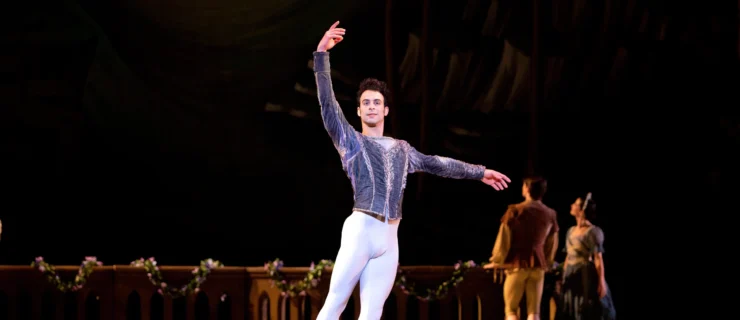 Arian Molina Soca, wearing a greay velvet tunic, white tights and gray ballet boots, stands in tendu derrierre effacé with his right leg extended and his right arm up. He looks out towards the audience with a small smile. Behind him upstage are two dancers in peasant costumes, standing casually along a low wall covered in floral garland. They face upstage, looking at the moonlight shining from the backdrop.