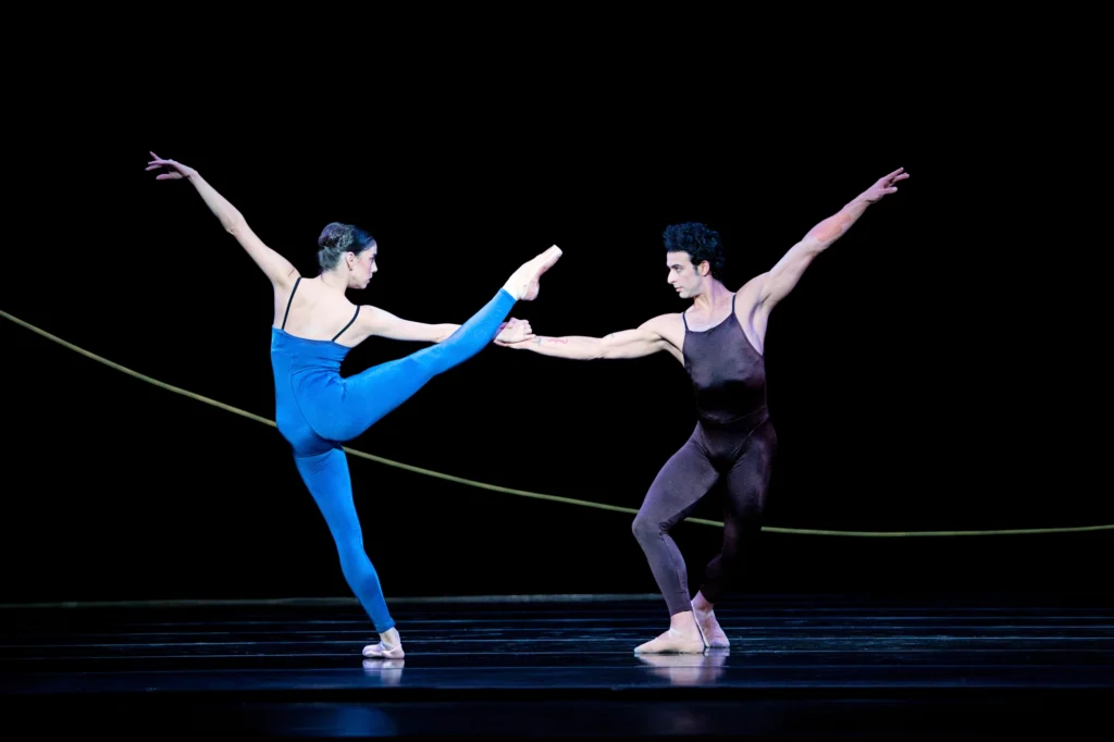 Dayesi Torriente and Arian Molina Soca dance a pas de deux onstage in front of a black backdrop wth a thin, curved green line towards the bottom. Torriente faces upstage and extends her right leg high to the side. She holds Soca's right hand with her own right hand and pulls him slightly as he promenades around her, his legs in plié. They each extend their left arms high to side. Torriente wears a blue unitard and pointe shoes, while Soca wears a dark purple unitard and tan ballet slippers.