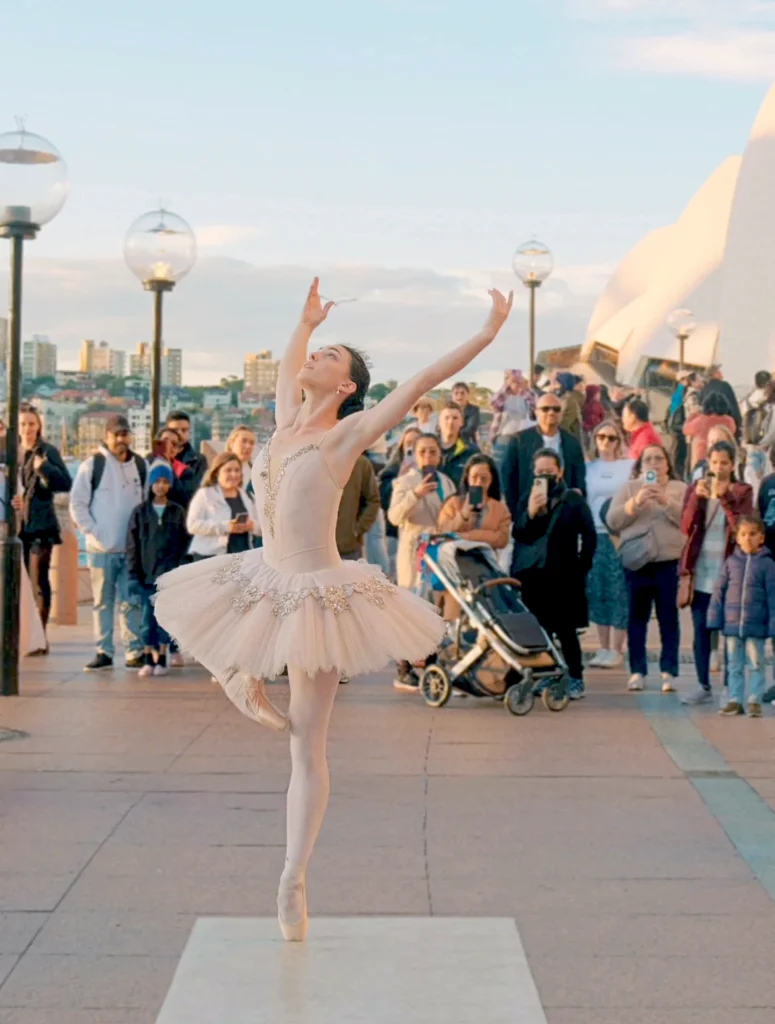 Bianca Carnovale, wearing a light pink tutu with a tiara, dances on pointe before a crowd on the street.
