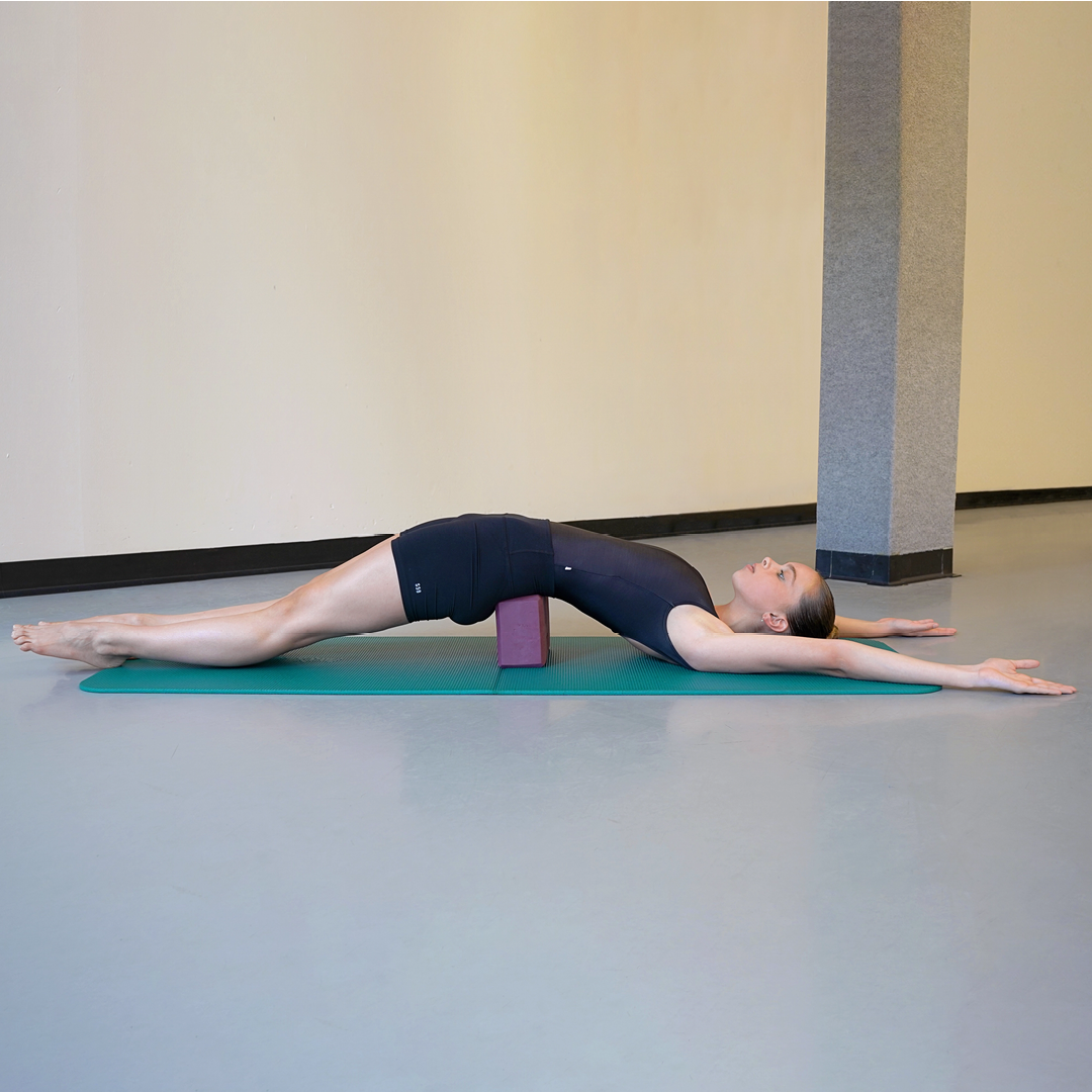 Model Stella MacDonald demonstrates the supported bridge pose with the arms and legs outstretched. Her hair is in a high bun and she is wearing black yoga attire. She is using one standard-sized, purple yoga block and a teal mat.