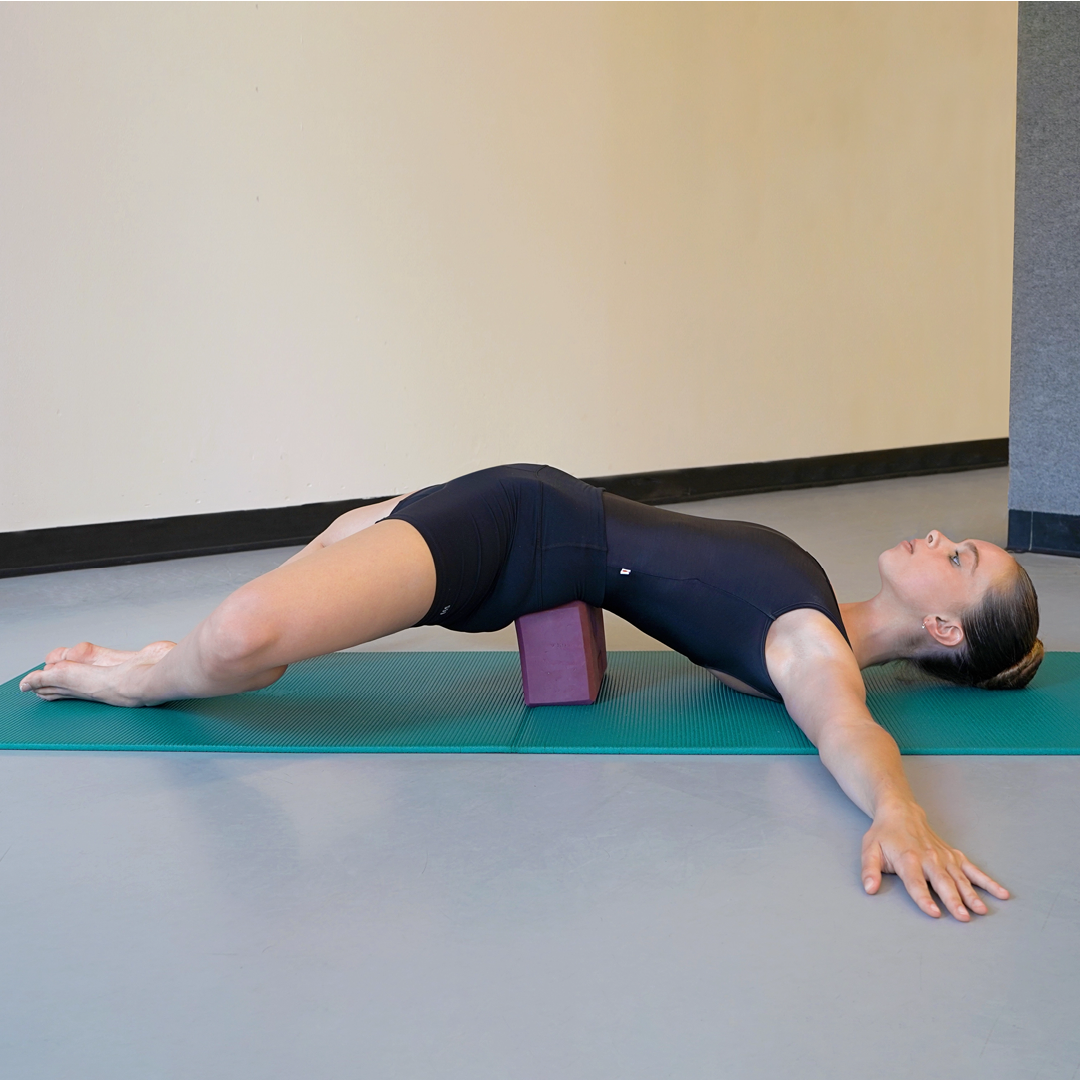 Model Stella MacDonald demonstrates the supported bridge pose with a butterfly variation. Her hair is in a high bun and she is wearing black yoga attire. She is using one standard-sized, purple yoga block and a teal mat.