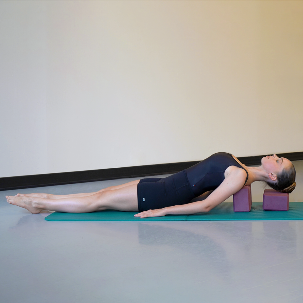 Model Stella MacDonald demonstrates the supported fish pose with the shoulder-supporting block turned vertically for higher elevation. Her hair is in a high bun and she is wearing black yoga attire. She is using two standard-sized, purple yoga blocks and a teal mat.