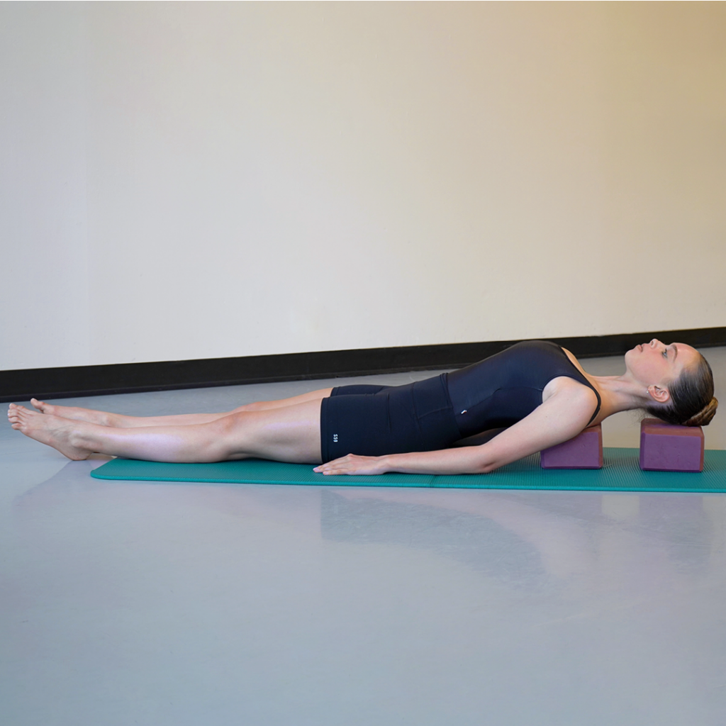 Model Stella MacDonald demonstrates the supported fish pose. Her hair is in a high bun and she is wearing black yoga attire. She is using two standard-sized, purple yoga blocks and a teal mat.