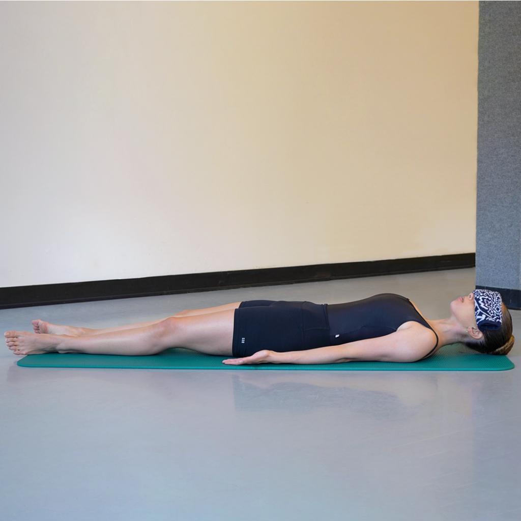 Model Stella MacDonald demonstrates savasana with a small towel covering her eyes. Her hair is in a high bun and she is wearing black yoga attire. She is using a teal yoga mat.
