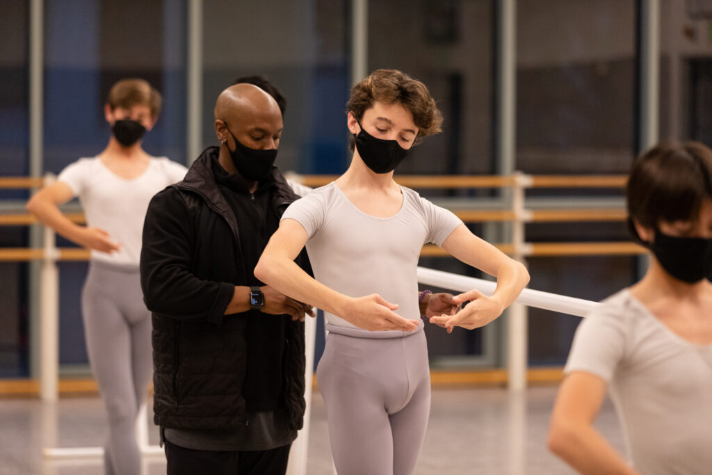 Jason Ambrose, wearing a black shirt, face mask, and puffy vest, corrects a male student during a ballet barre. The young student is shown from the thighs up holding his arms in first postition and wearing gray tights, a white T-shirt and black face mask. Ambrose moves his hands towards his waist to correct the boy's placement.