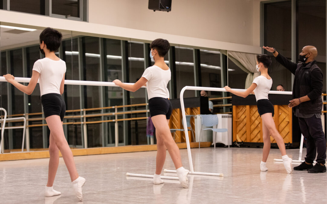Three young boys stand with their left leg in tendu derriere facing the barre in front of a mirror in a large dance studio. They wear white T-shirts, black shorts, white socks and ballet slippers and face masks. Jason Ambrose, wearing black athletic clothing, dance sneakers, and a black face mask, stands at the end of the barre with his hand above one boy's head, as if instructing him to stand taller.