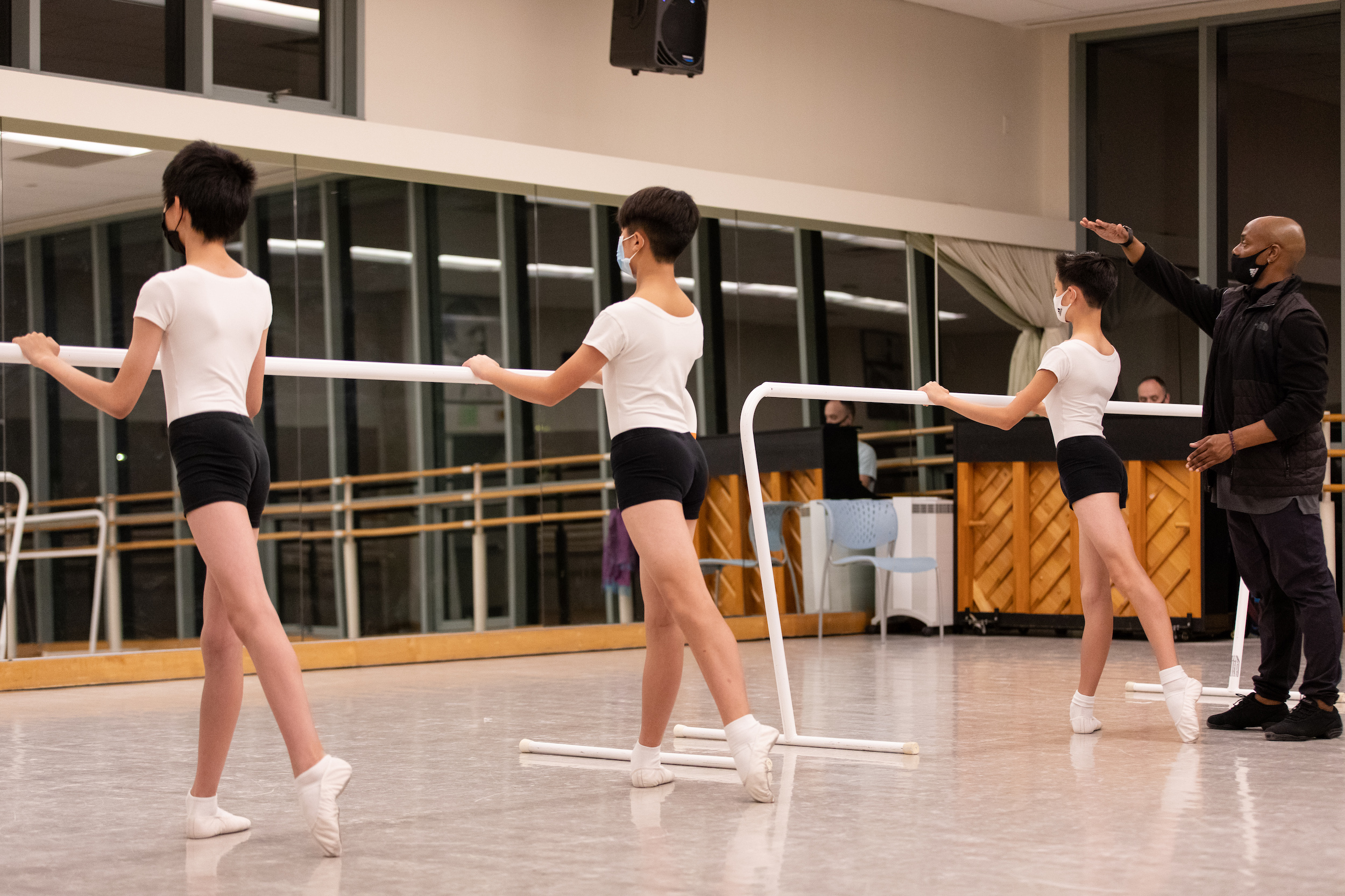 Three young boys stand with their left leg in tendu derriere facing the barre in front of a mirror in a large dance studio. They wear white T-shirts, black shorts, white socks and ballet slippers and face masks. Jason Ambrose, wearing black athletic clothing, dance sneakers, and a black face mask, stands at the end of the barre with his hand above one boy's head, as if instructing him to stand taller.