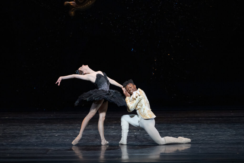 Maria Kochetkova and Siphesihle November pose during a performance of Swan Lake. Kochetkova, wearing a black tutu and tiara, stands in tendu derriere facing November and arches her body back, her right arm reaching long alongside her head. November kneels and clasps her left and, leaning his cheek against it, closing his eyes and smiling. He wears a white and gold tunic, white tights and white boots.
