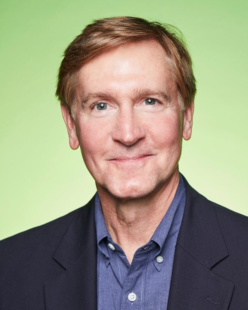 Tim O'Keefe, shown shoulders-up, poses for a portrait photo in front of a light green backdrop. He smiles playfully with his mouth closed.