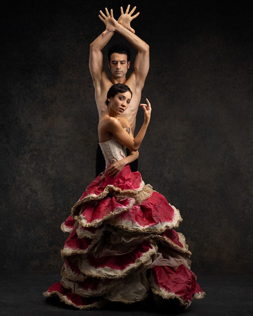Nayara Lopes and Arian Molina Soca pose together for a photo promoting "Carmen." Lopez wears a long, layered scarlet skirt with gold trimming and a gold bodice; she's turned profile and looks into the camera seductively. Molina Soca stands directly behind her, only his chest-up visible, and he raises his arms above his head, hands crossed at the wrist, with passion.