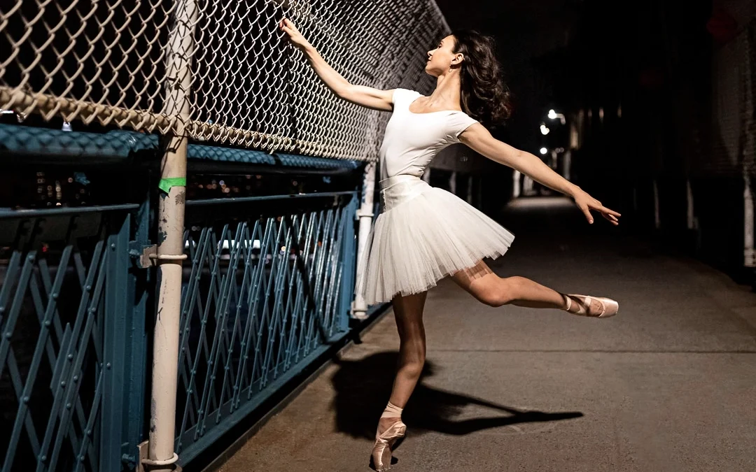 Camila Rodrigues poses in a low attitude effacé on pointe outdoors at nighttime, grasping a chain-link fence with her right hand and stretching her left arm out low and behind her. She wears a white leotard and tutu, and pointe shoes, and looks over her right hand.