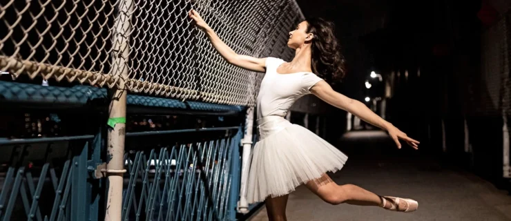 Camila Rodrigues poses in a low attitude effacé on pointe outdoors at nighttime, grasping a chain-link fence with her right hand and stretching her left arm out low and behind her. She wears a white leotard and tutu, and pointe shoes, and looks over her right hand.