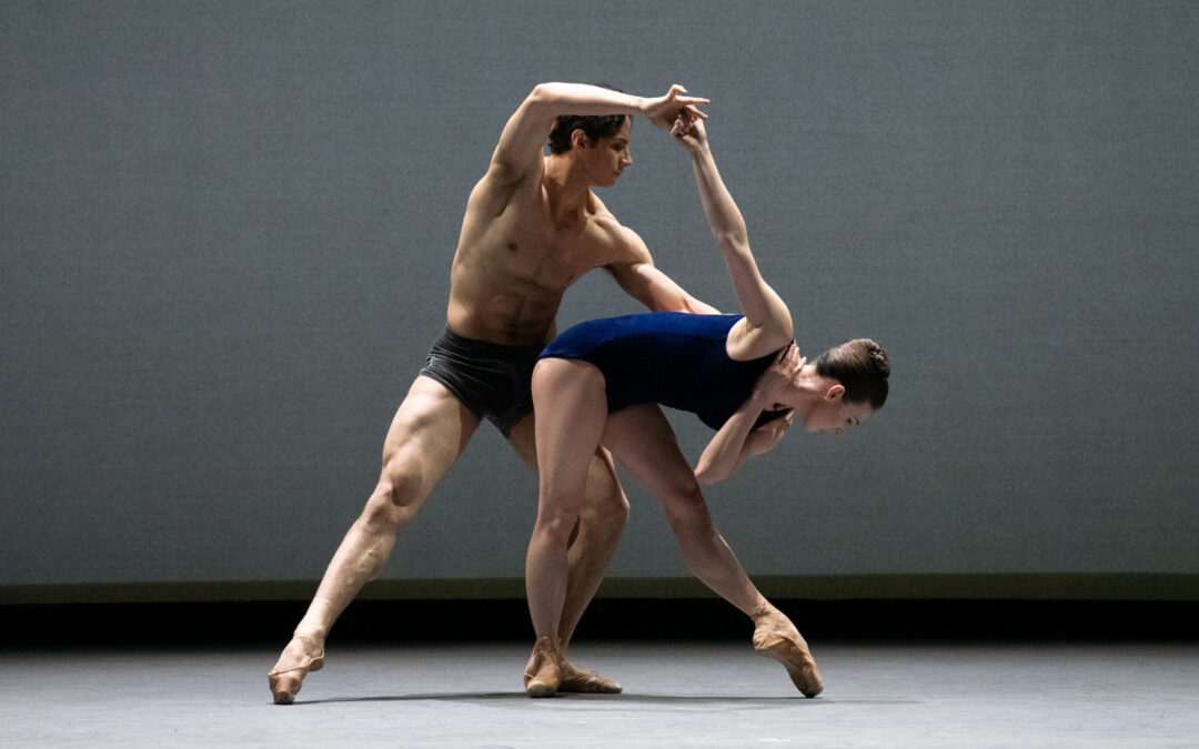 Tiler Peck and Roman Mejia dance together onstage in front of a gray backdrop. Peck wears a deep blue leotard, and Mejia dark blue dance shorts. Peck bends over in a stylized tendu, and Mejia lunges in a parallel tendu, holding her right arm up and supporting her waist.
