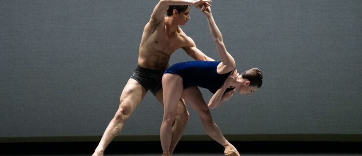 Tiler Peck and Roman Mejia dance together onstage in front of a gray backdrop. Peck wears a deep blue leotard, and Mejia dark blue dance shorts. Peck bends over in a stylized tendu, and Mejia lunges in a parallel tendu, holding her right arm up and supporting her waist.