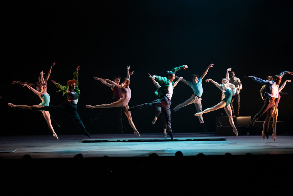 Lit in a spotlight, a large group of dancers suspend in mid-air in an arabesque saute.