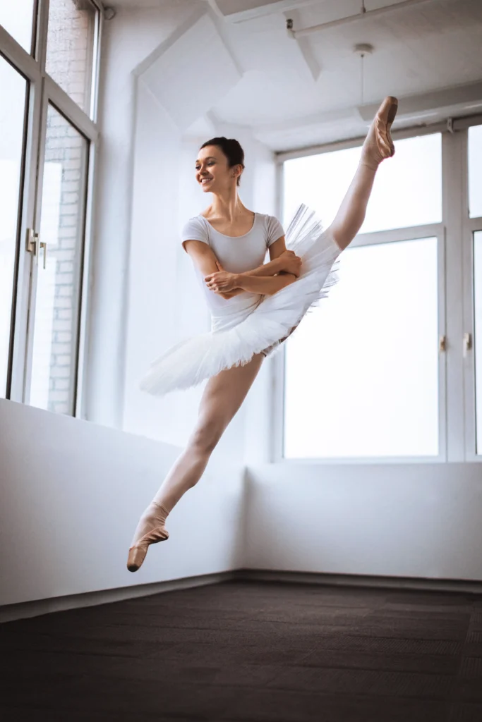 Camila Rodrigues jumps up, splitting her legs in a tilted position so that her left leg is high. She crosses her arms in front of her and looks over her right shoulder, smiling wide. She wears a white leotard and white tutu, pink tights and pink pointe shoes.