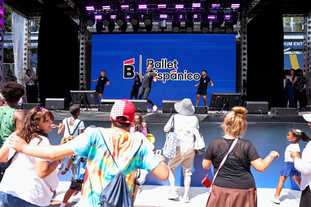 A group of dancers in black T-shirts and shorts or leggings dances on an outdoor stage while onlookers watch and dance along with them. The dancers perform in front of a lit blue backdrop with the words Ballet Hispánico on it.