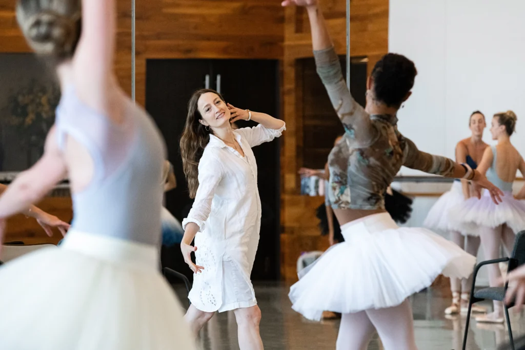 Julie Kent, wearing a white, long-sleeved dress, is shown facing a group of dancers. She touches her left hand to her face and tilts her head to the side, coaching them to use more épualment as they pose in a position. The female dancers wear leotards, tights and practice tutus and are mstly shown from the back. A few other dancers stand to the side, observing and talking.