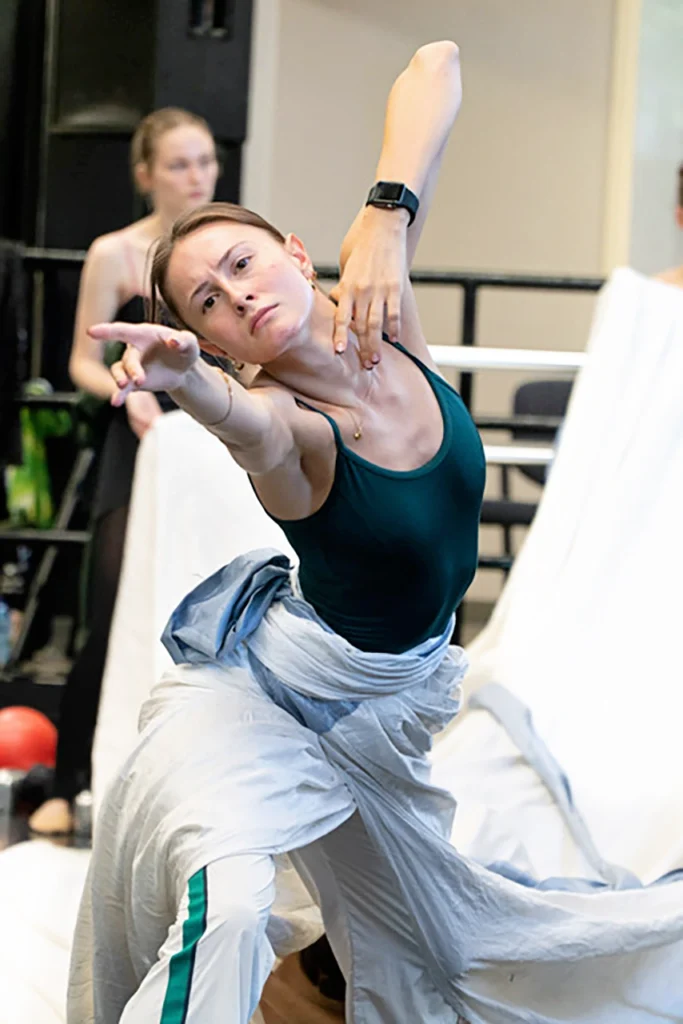 A close-up of a female dancer rehearsing in a studio. She wears a long, white fabric skirt that trails behind her and rests on a prop in the background. She reaches toward the camera with a determined, serious look, her body twisting up and her left arm bent so her left fingers touch her collarbone lightly.