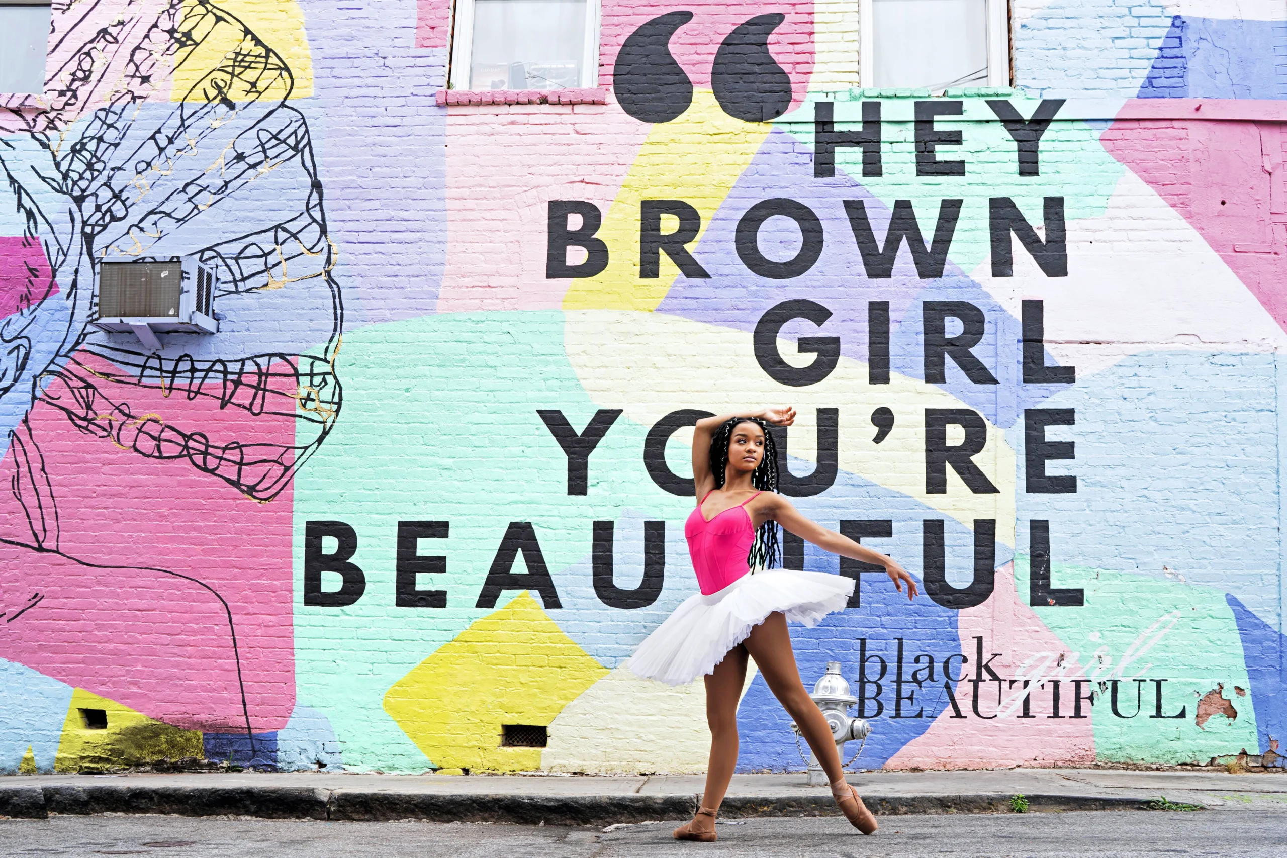 A young Black dancer poses in front of a colorful mural that says "Hey Brown Girl, You're Beautiful." She wears a hot pink leotard and a white tutu, with flesh-colored pointe shoes. She poses in tendu derriere, her torso twisted toward the camera. Her right arm drapes over her head, and her left extends in a low allongé behind her.