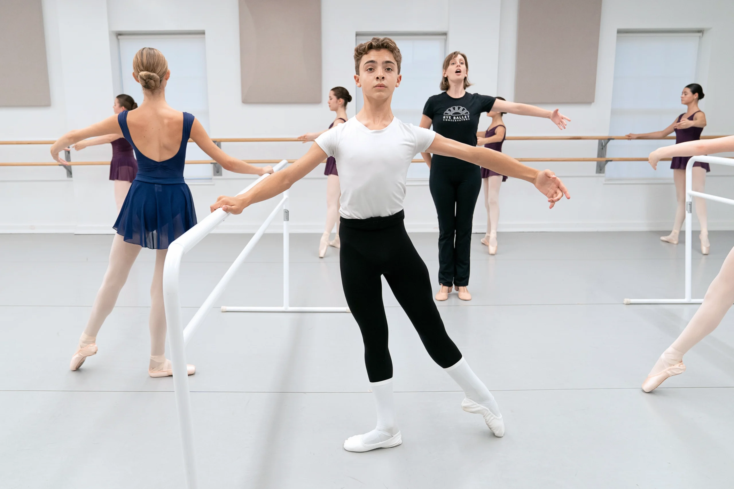 Thirteen-year-old Matthew Fontana, a ballet student, takes ballet class at a dance studio. He holds onto a portable barre with his right hand and does a tendu a la seconde with his left leg. He wears a white t-shirt, black tights, white socks and white ballet slippers. He is surrounded by female classmates who do the same exercise and how wear blue or purple leotards, pink tights, and pink pointe shoes. Their teacher, in a black t-shirt and yoga pants, stands among them with her feet in parallel and her left arm out in second position, talking to them as they dance.