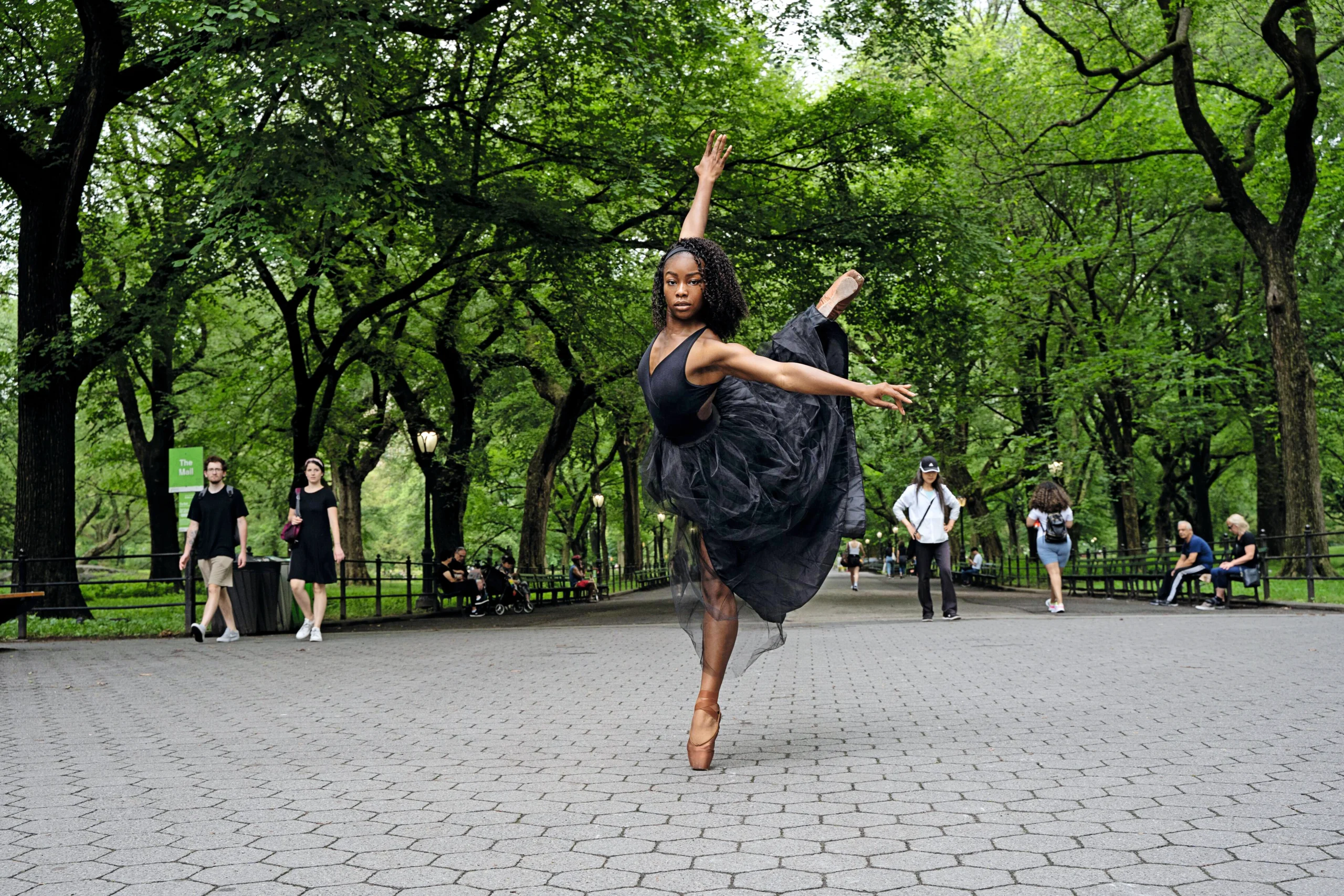 Outside in a public park, a young Black ballerina poses in a dramatic attitude derriere en croise. She wears a black dance dress and flesh-colored pointe shoes.