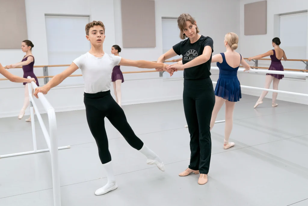 Thirteen-year-old Matthew Fontana, a ballet student, takes ballet class at a dance studio. He holds onto a portable barre with his right hand and does a degagé a la seconde with his left leg. He wears a white t-shirt, black tights, white socks and white ballet slippers. His teacher, Ashlee Knapp, shapes his left arm; they both look into the mirror, whicb is out of view. They are surrounded by female classmates who do the same exercise and wear blue or purple leotards, pink tights, and pink pointe shoes. Knapp a black t-shirt and yoga pants.