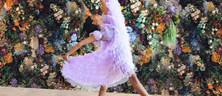 In front of a colorful floral backdrop, a young Black dancer poses for a photo wearing a long, ruffly light purple dress. She swishes the dress up with her hands, leaning back slightly in a controlled fall off-pointe forward.