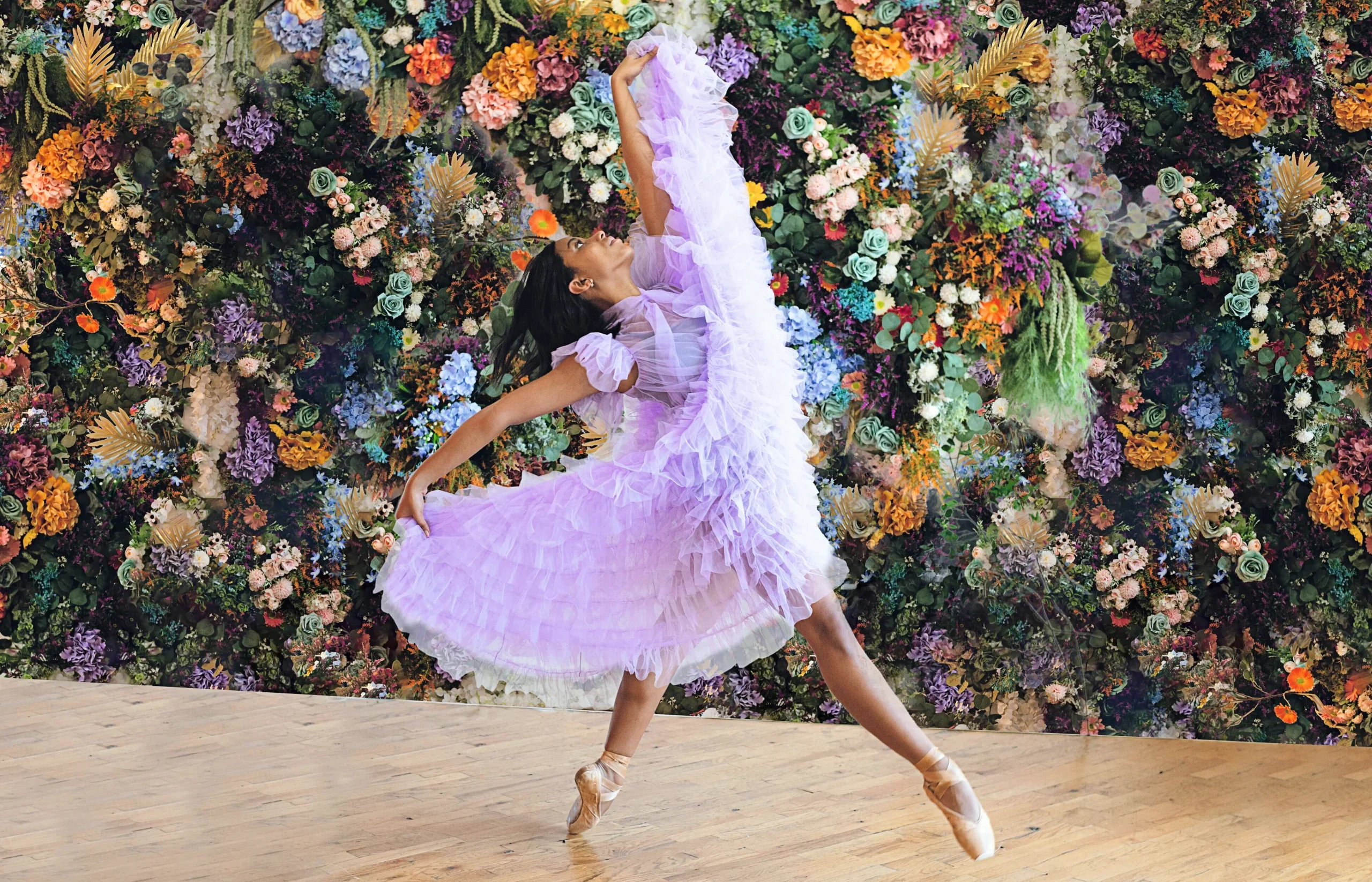 In front of a colorful floral backdrop, a young Black dancer poses for a photo wearing a long, ruffly light purple dress. She swishes the dress up with her hands, leaning back slightly in a controlled fall off-pointe forward.