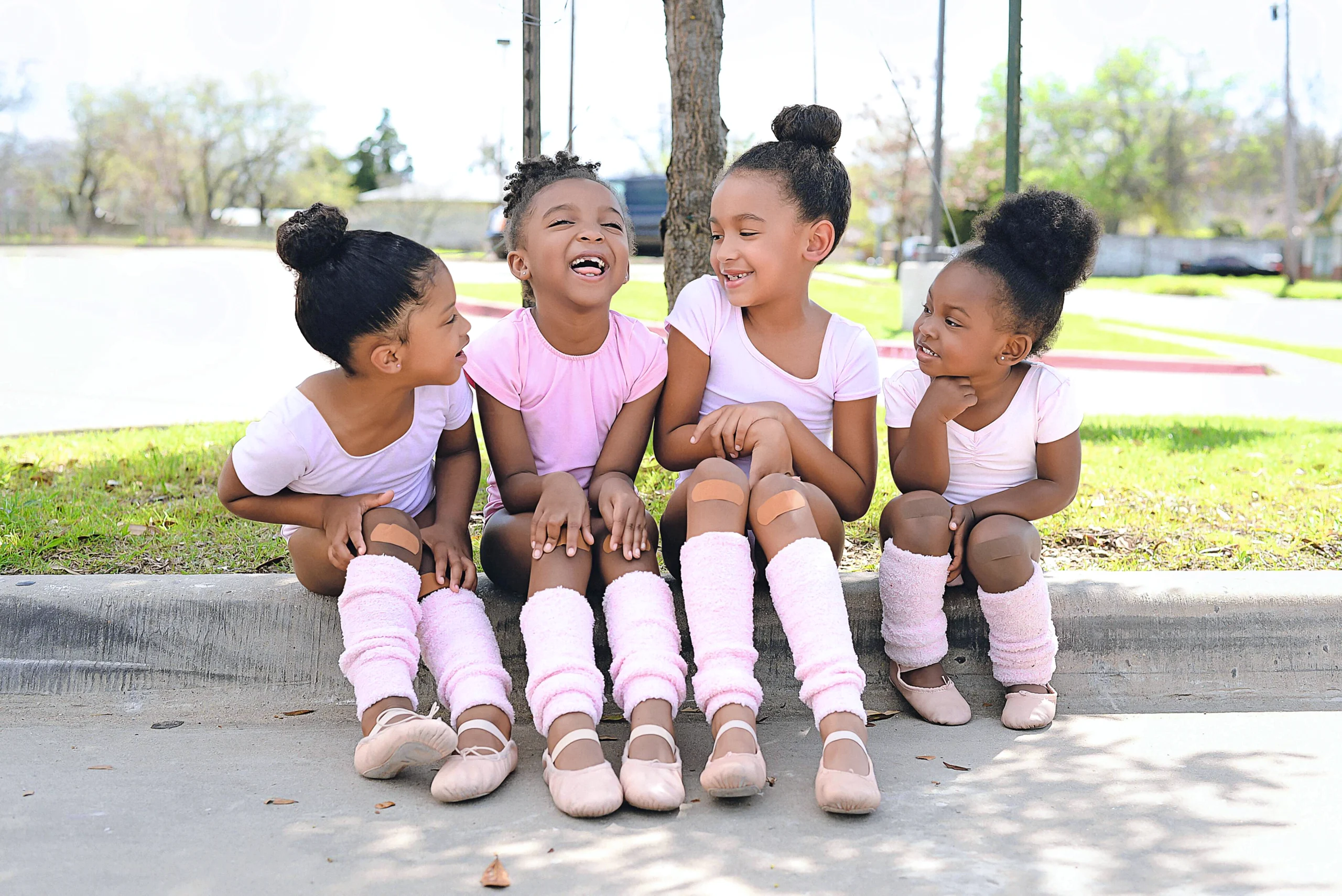 Outside on a sunny day, four young Black ballerinas (approximately age 5) sit together on a curb and laugh. They all wear light pink cap-sleeve leotards and pink leg warmers.