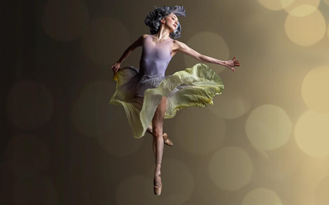 In front of an ombré gold background with large spots of light, a female dancer is suspended mid-air in a retiré jump, her silver hair flying around her face and her arms flung to the sides. She wears a light and flowy skirt with light purple and light green sections.