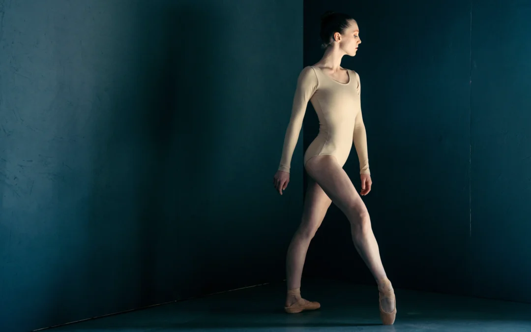 A ballerina poses mysteriously in front of a dark gray backdrop. She wears a long-sleeved nude leotard and nude pointe shoes and poses with her right leg in forced arch, croisé devant. She turns her head away from the camera and is lit with dark lighting.