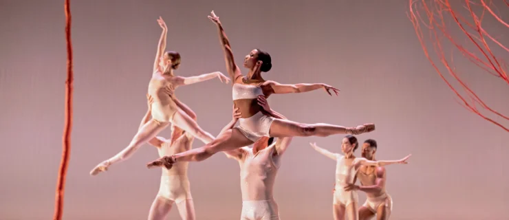Rachael Parini dos a large sissone, her legs split, as her male partner holds her by the waist and lifts her high into the air onstage. Two other couples do the same step in the background. The women wear light-colored shorts, bralette tops, and pointe shoes, and the men are shirtless with light-colored shorts. They dance in front of a taupe backdrop and a few orange trees.