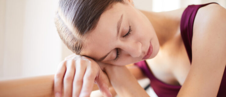 Exhausted young female dancer with closed eyes tired from ballet training leaning on barre and sleeping in studio
