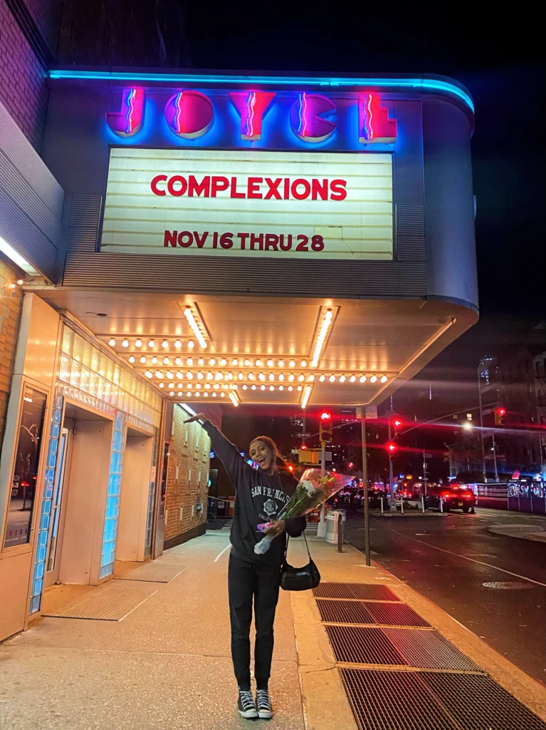 Jasmine Robinson, wearing a dark sweatshirt and sweatpants and tennis shoes, stands outside under The Joyce Theater's marquis, holding a bouquet of flowers and lifting her right arm triumphantly. The marquis sign says Complexions in red lettering.