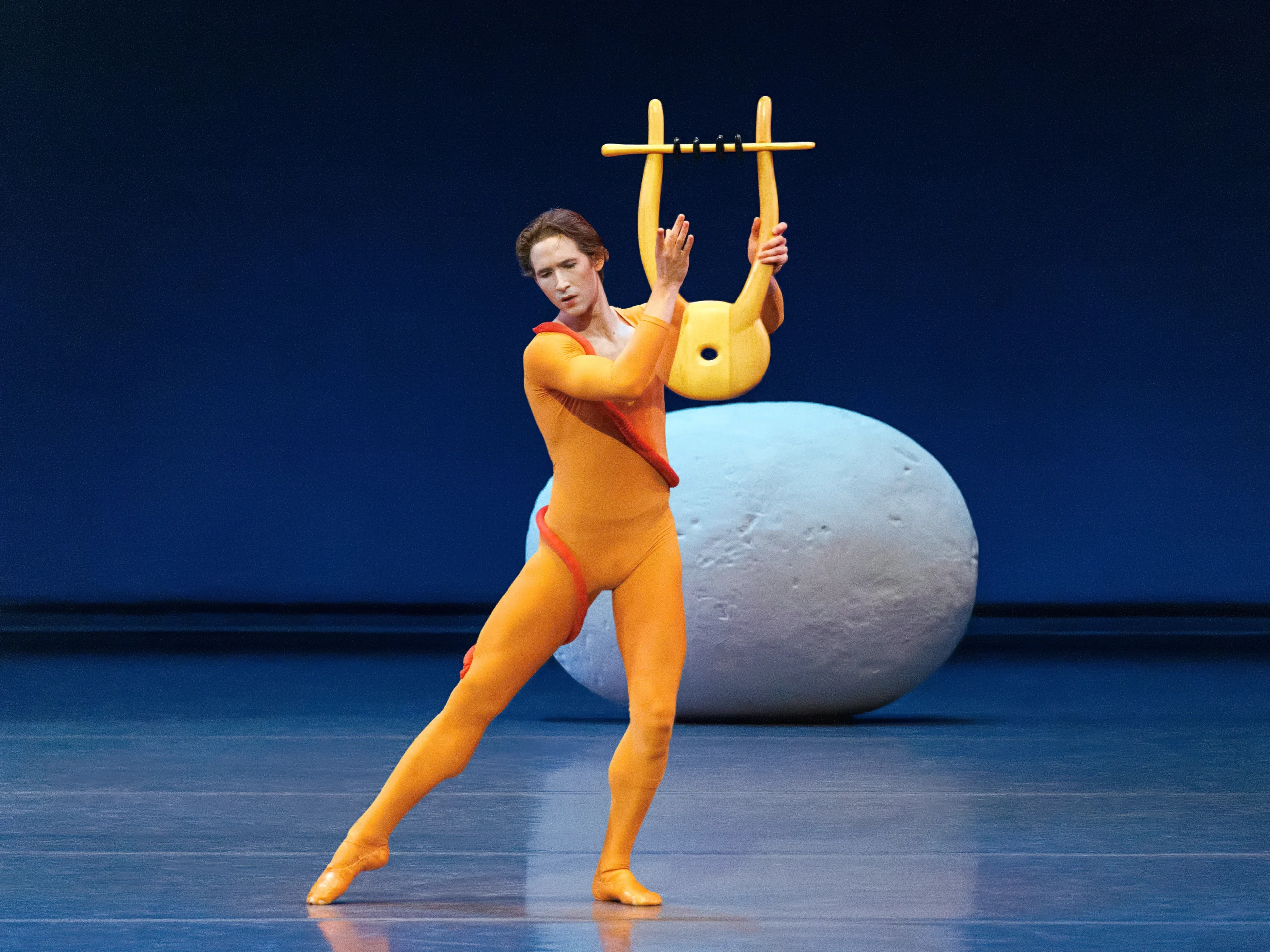 Joseph Gordon dances in "Orpheus" as the title role. He wears a bright yellow-orange unitard and holds a large lyre prop. A rounded rock prop rests behind him.
