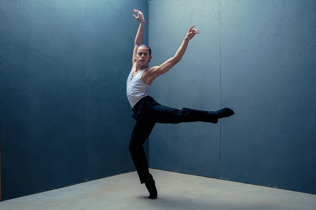 In front of a dark blue backdrop, a male dancer poses in a forced arch attitude derriere, his arms flying upward gently.