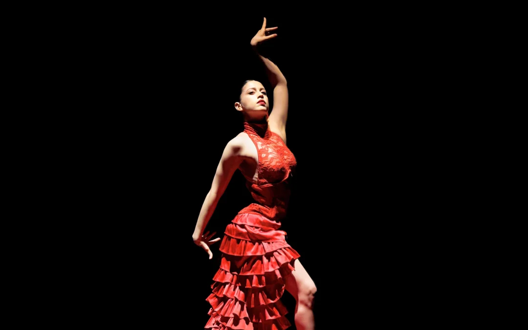 Amanda del Valle, wearing a bright red flamenco-style dress, stands onstage in a pool of light, surrounded by darkness. She lifts her left arm up above her head, breaking at the wrist, and keeps her right arm down straight behind her making the same wrist shape. She looks out confidently towards the audience.
