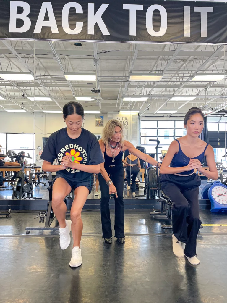 Lisa Giannone, standing behind and between dancers WanTing Zhao and SunMin Lee, instructs them during an exercise. Zhao and Lee are in a high, one-legged squat with their hands clasped and elbows bent.