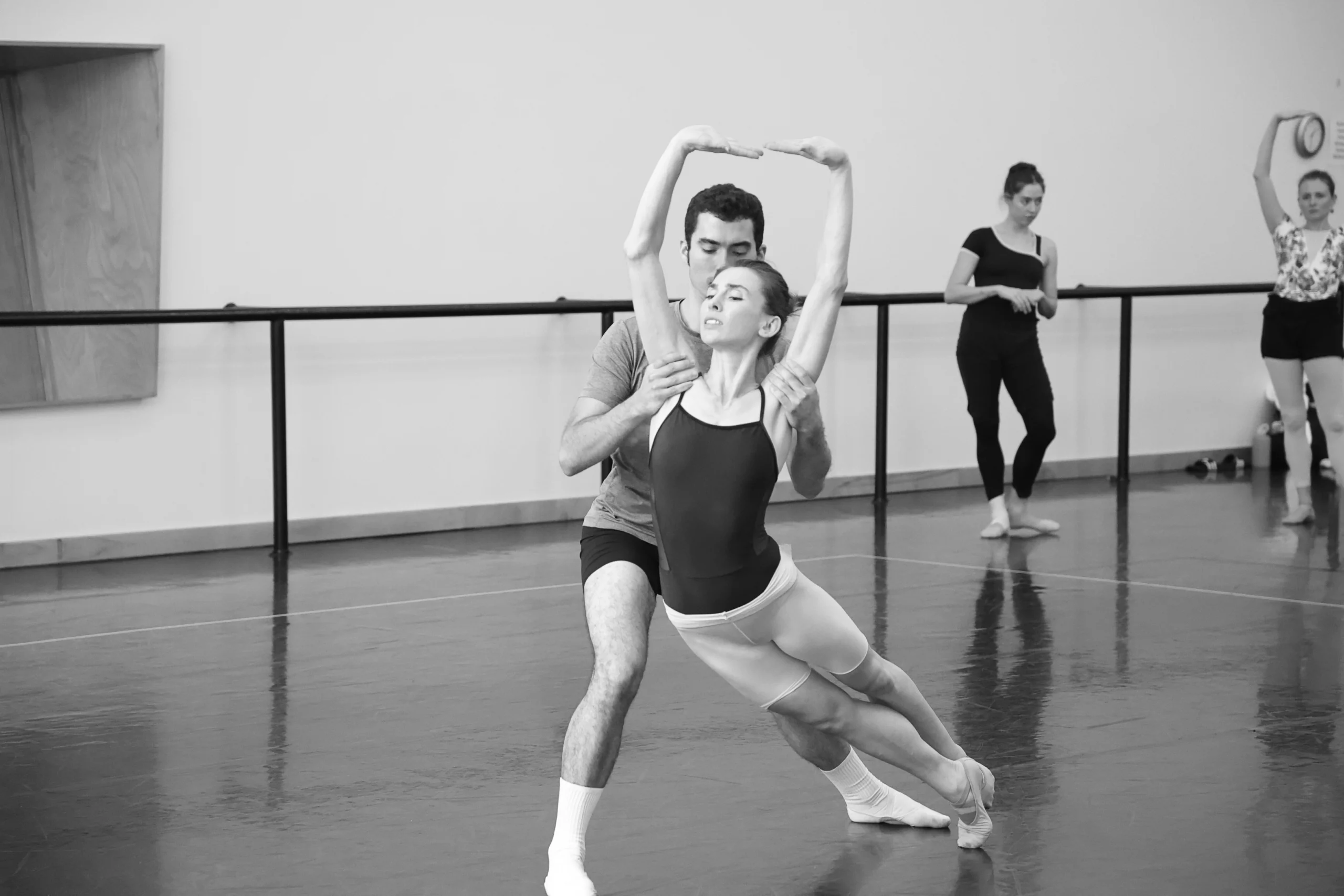 In this black and white photo, a male dancer lunges on his right leg and holds a female dancer by the upper arms as she leans into her hips in profile, her legs crossed in sus-sous. She holds her arms in high fifth with her hands flexed and looks towards the right. The ballerina wears a dark spaghetti strap leotard, light-colored tights cut off at the thighs and ballet slippers. The male dancer wears dark shorts, a T-shirt, whte socks and ballet shoes. Two other female dancers practice in the back of the dance studio.