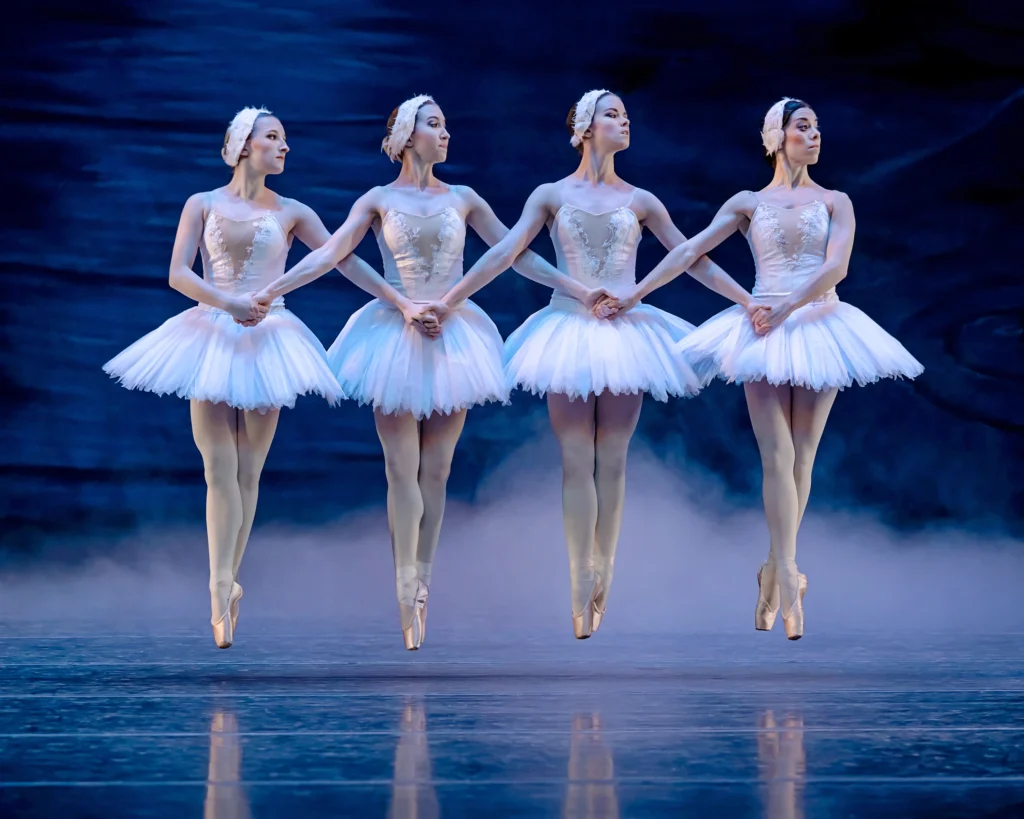 A quartet of female ballerinas link hands and perform an entrechat quatre during a performance of Swan Lake. They all wear white tutus, white feathered headpieces, pink tights and pink pointe shoes. The dance in front of a dark lake-scene backdrop with fog billowing from the ground.