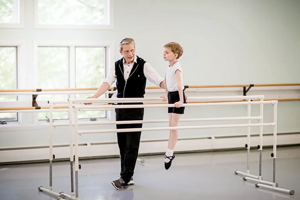 In a large dance studio, Marc Pielbergerstands next to three portable ballet barres and coaches a young boy as he lifts himself up between two barres, using his arms to push himself up and pointing his feet off the floor. The little boy wears a white t-shirt, black shorts, white socks and black ballet slippers. Pielberger wears black pants and sneakers, a white button down shirt with a black sleeveless zip-up vest over it.